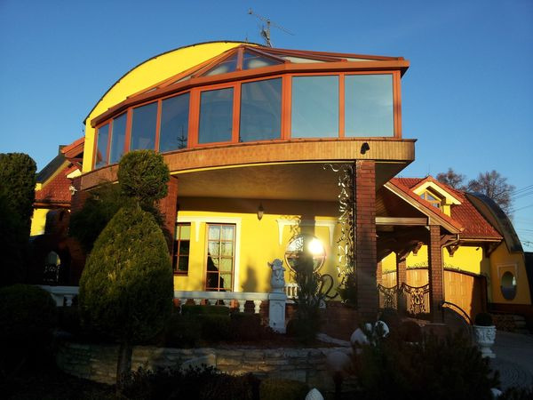 Żółty dom z zabudowanym balkonem z drewnianą ramą okienną i okrągłym oknem. Wokół domu ozdobne krzewy i drzewa.