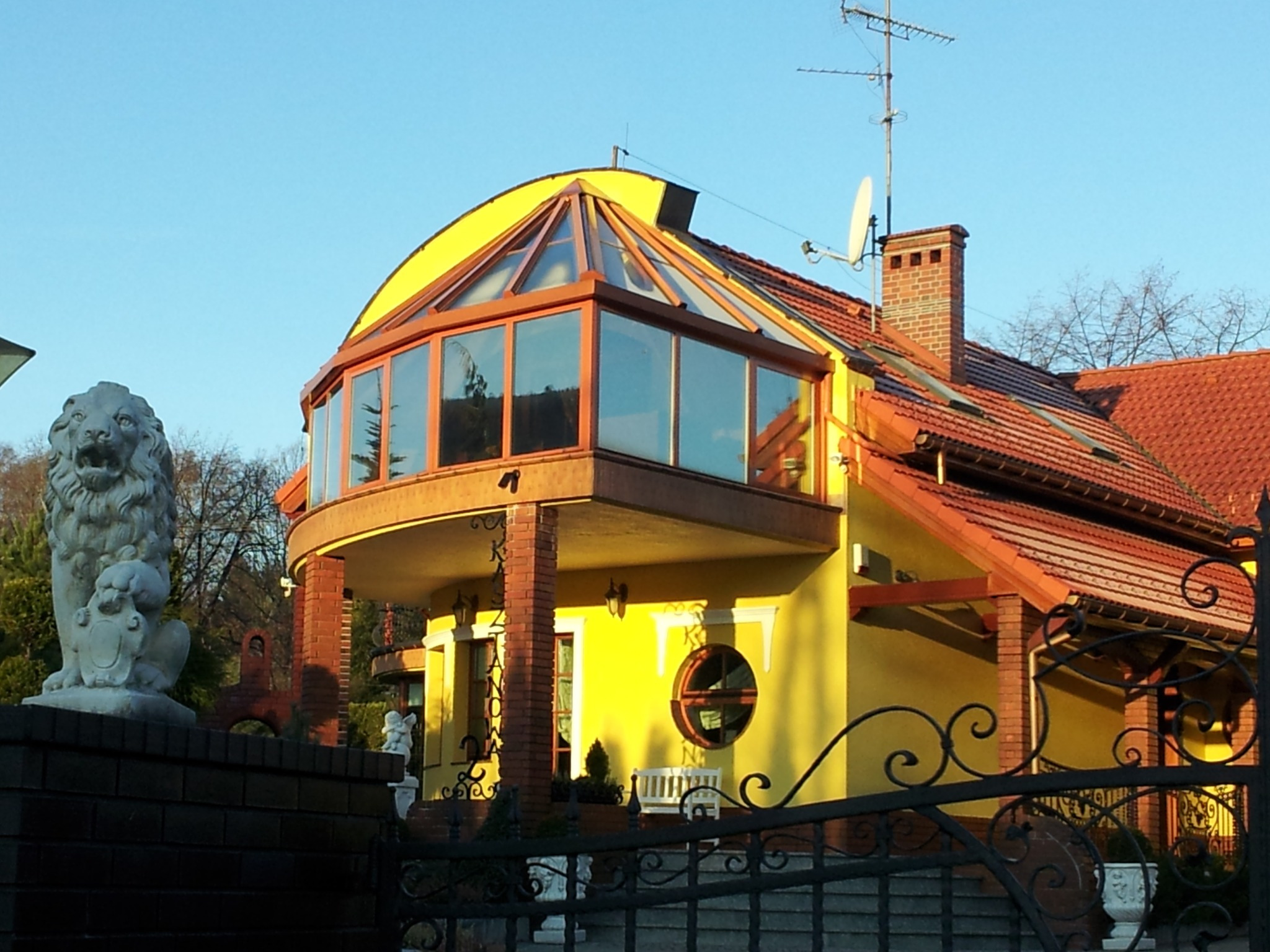 Żółty dom z zabudowanym balkonem w stylu oranżerii, widok zza ozdobnego ogrodzenia. Na pierwszym planie rzeźba lwa.