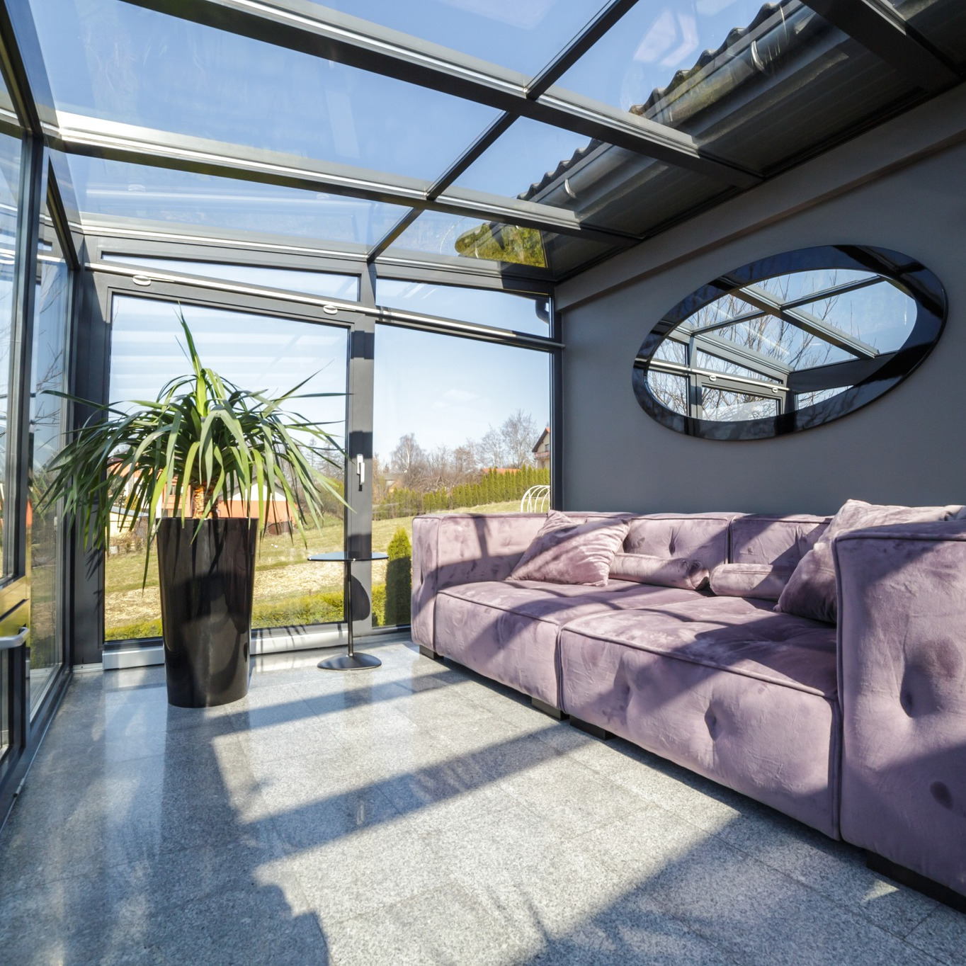 Szklana zabudowa balkonu z metalową konstrukcją, widok na ogród. Wnętrze urządzone w nowoczesnym stylu z sofą w kolorze pudrowego różu i dużym lustrem.