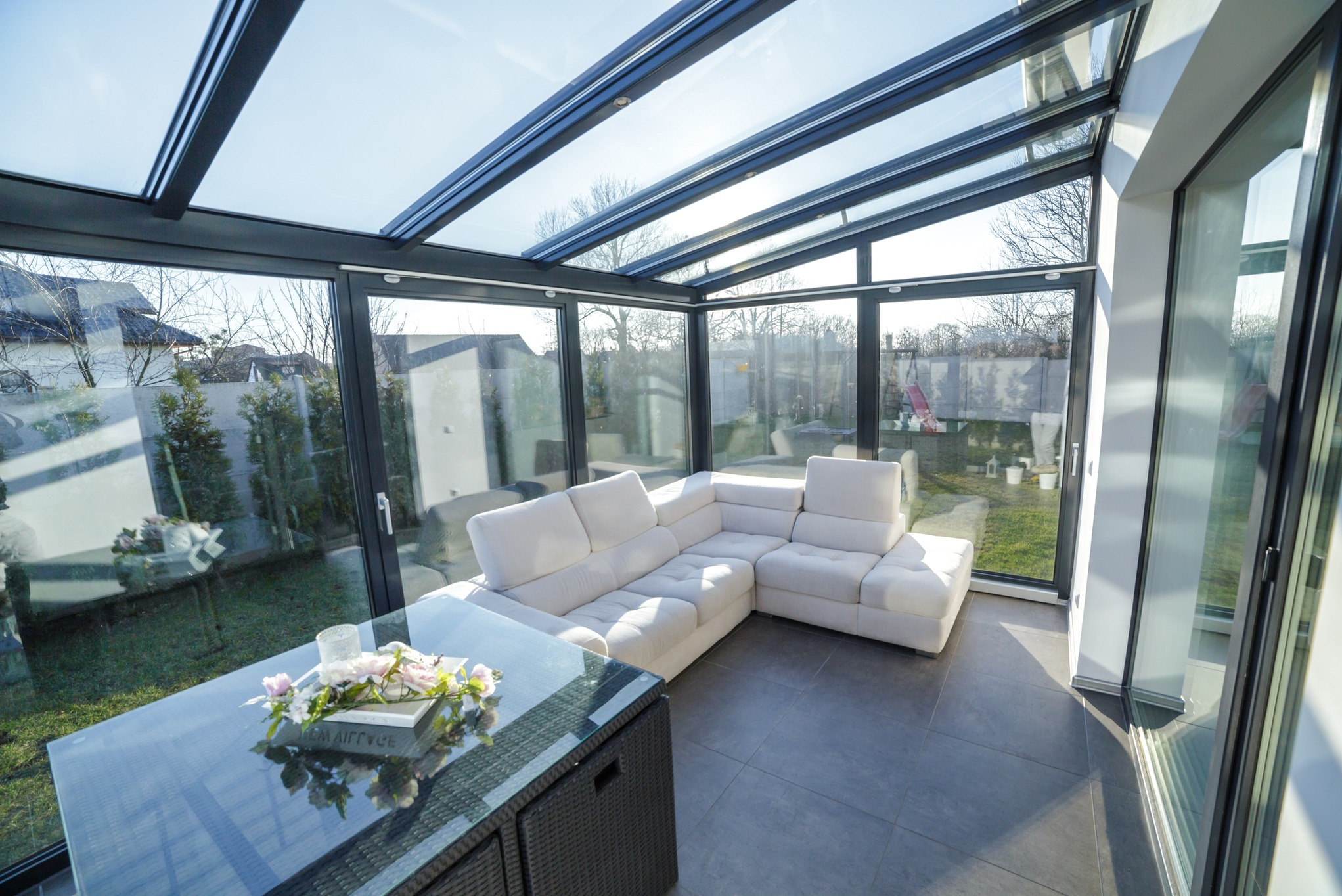 Nowoczesna zabudowa balkonu z metalową konstrukcją i szklanym dachem, wewnątrz biała sofa i stolik z dekoracją kwiatową. Widok na ogród.