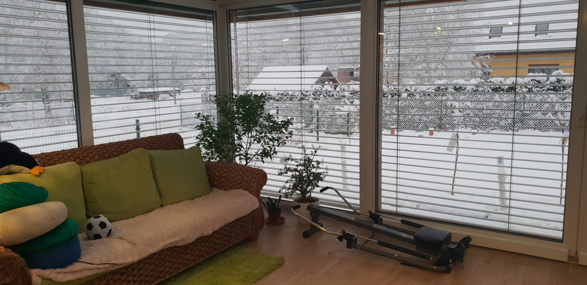 Zabudowany balkon z widokiem na zimowy krajobraz. Wnętrze z wiklinową sofą, zielonymi poduszkami i roślinami w doniczkach. Na podłodze leży sprzęt do ćwiczeń.
