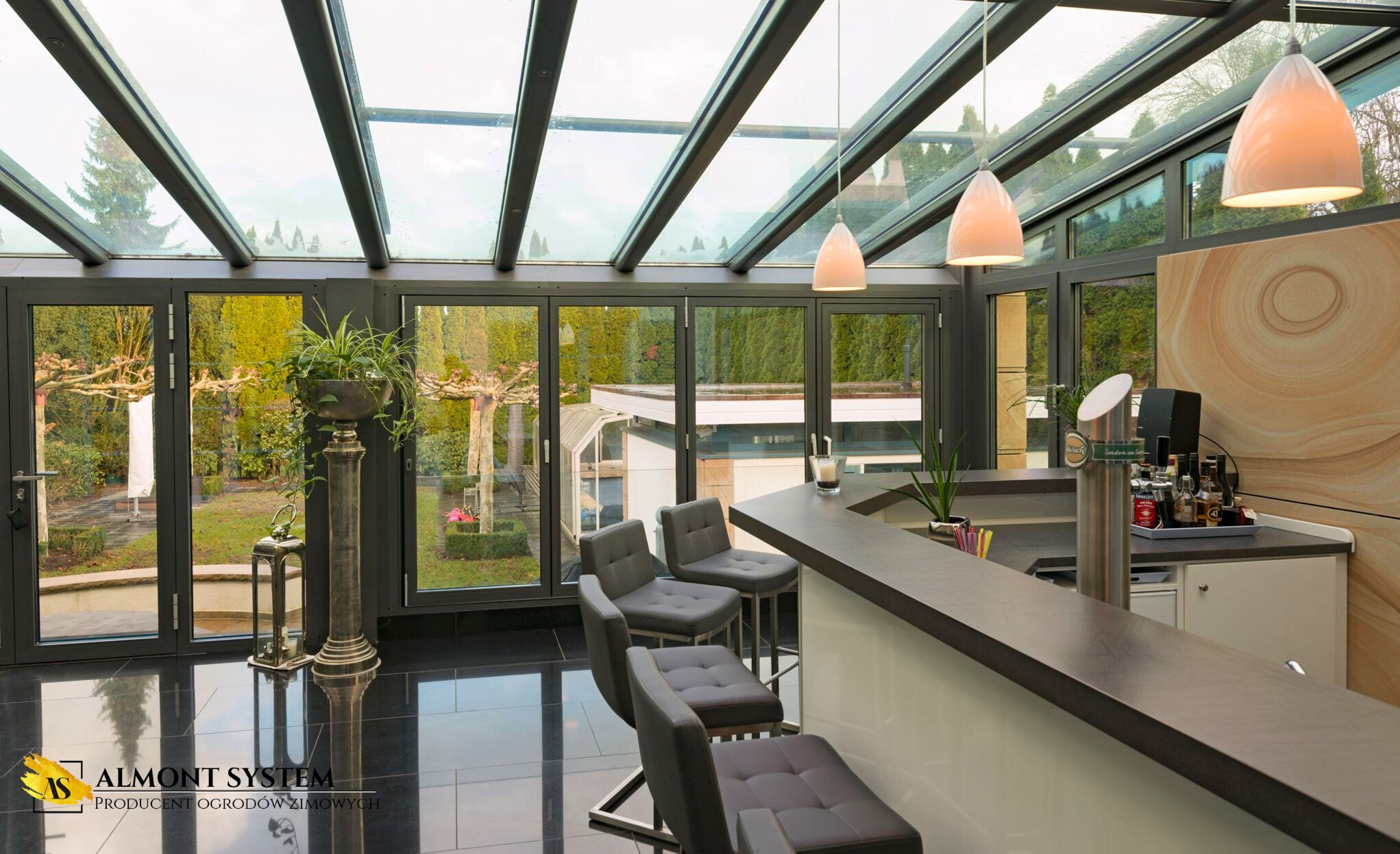 Zimowy ogród z metalową konstrukcją i szklanym dachem, wyposażony w bar z krzesłami. Widok na ogród przez duże okna w grafitowych ramach.