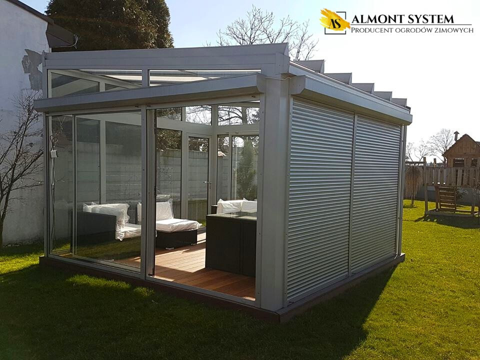 Metalowa pergola z roletami i szklanymi ścianami, wyposażona w meble ogrodowe, na tle zadbanego trawnika i budynku. Słoneczny dzień.