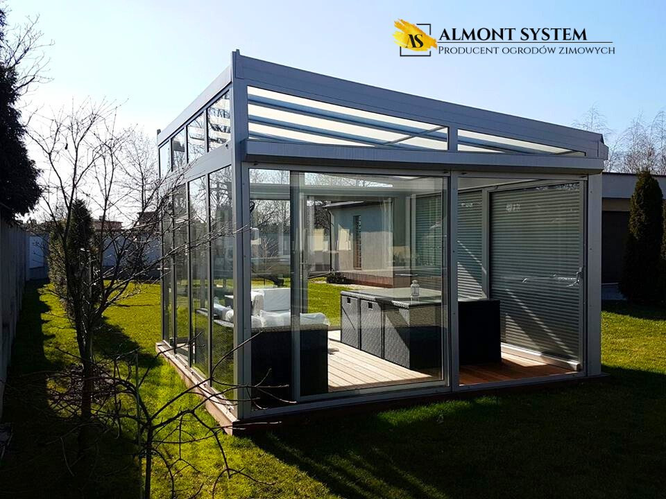 Szklana, nowoczesna pergola na trawniku, z meblami w środku. Widoczne rolety i przeszklone ściany. Słoneczny dzień.