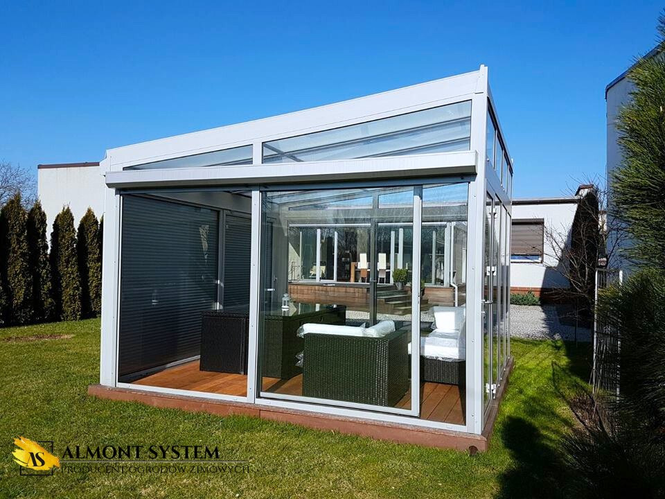 Szklana pergola z metalową konstrukcją na drewnianym podeście, zacieniona roletami, z widocznym wyposażeniem wnętrza i zielonym otoczeniem.