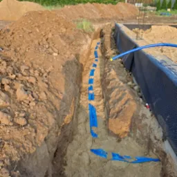 Wykop z ułożonymi niebieskimi rurami instalacji w piasku, zabezpieczonymi taśmą ostrzegawczą. Fundament budynku w tle. Widok z góry na instalację.