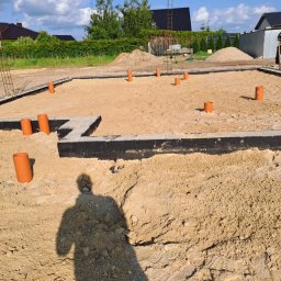 Jarek Plewa - Przygotowanie fundamentów pod budynek z wystającymi rurami kanalizacyjnymi, zabezpieczone hydroizolacją, teren wyrównany piaskiem, widoczny cień osoby.