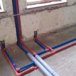 Instalacja ogrzewania podłogowego w trakcie montażu: niebieskie i czerwone rury ułożone na surowym betonie, wychodzące ze ściany, widoczne okno w tle.