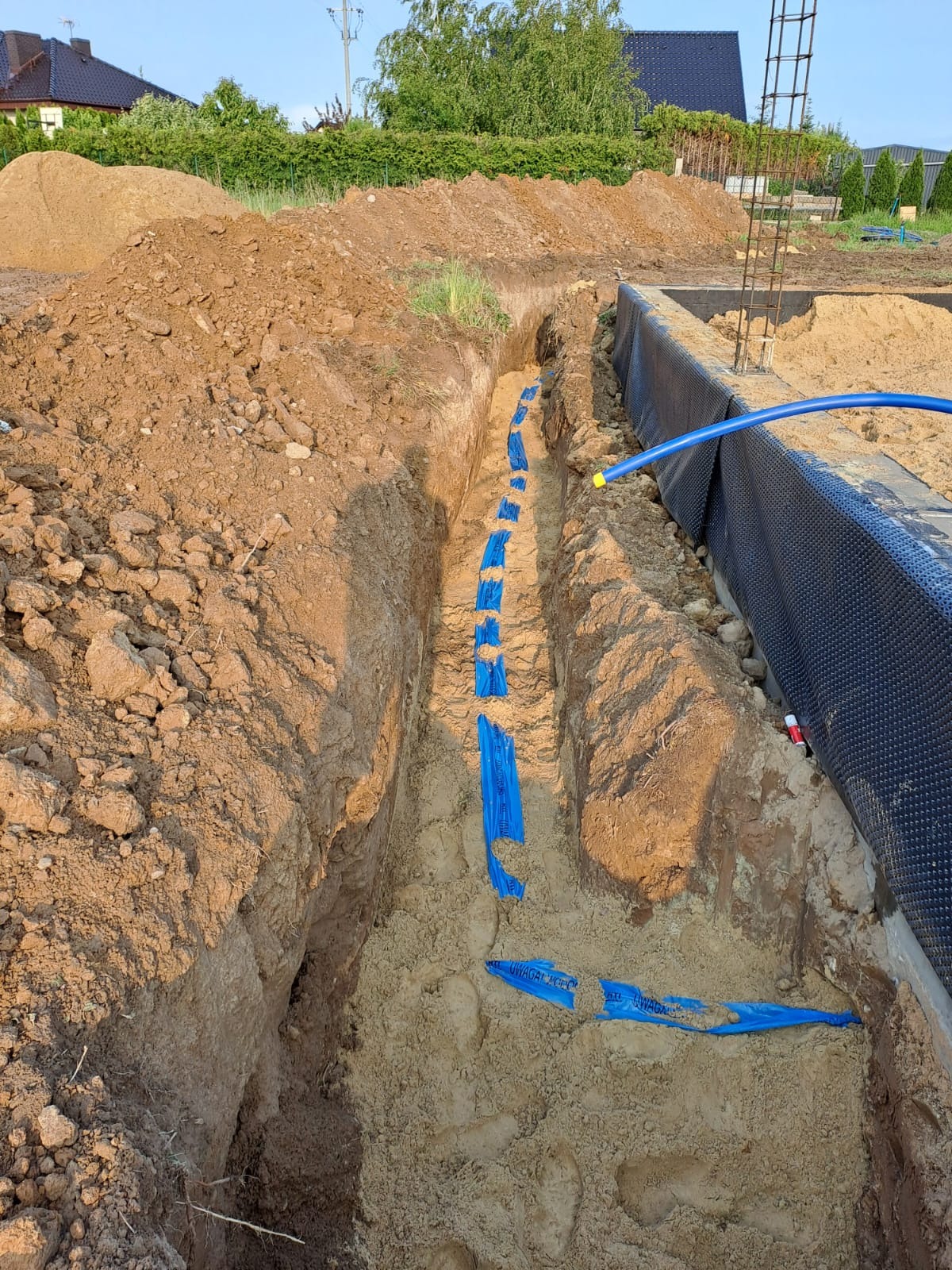 Wykop z ułożonymi niebieskimi rurami instalacji w piasku, zabezpieczonymi taśmą ostrzegawczą. Fundament budynku w tle. Widok z góry na instalację.