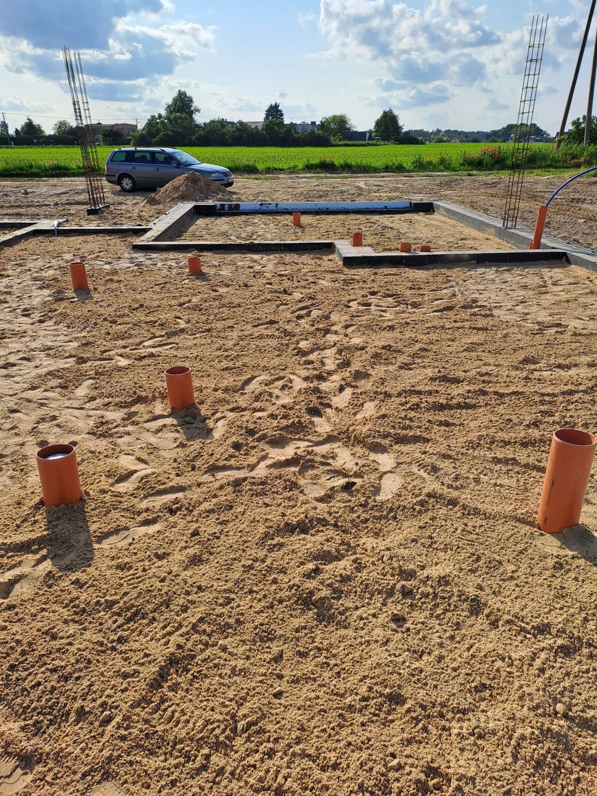 Fundamenty domu jednorodzinnego z wystającymi rurami, na tle pola i błękitnego nieba z chmurami. Widoczne zbrojenia i szalunki.