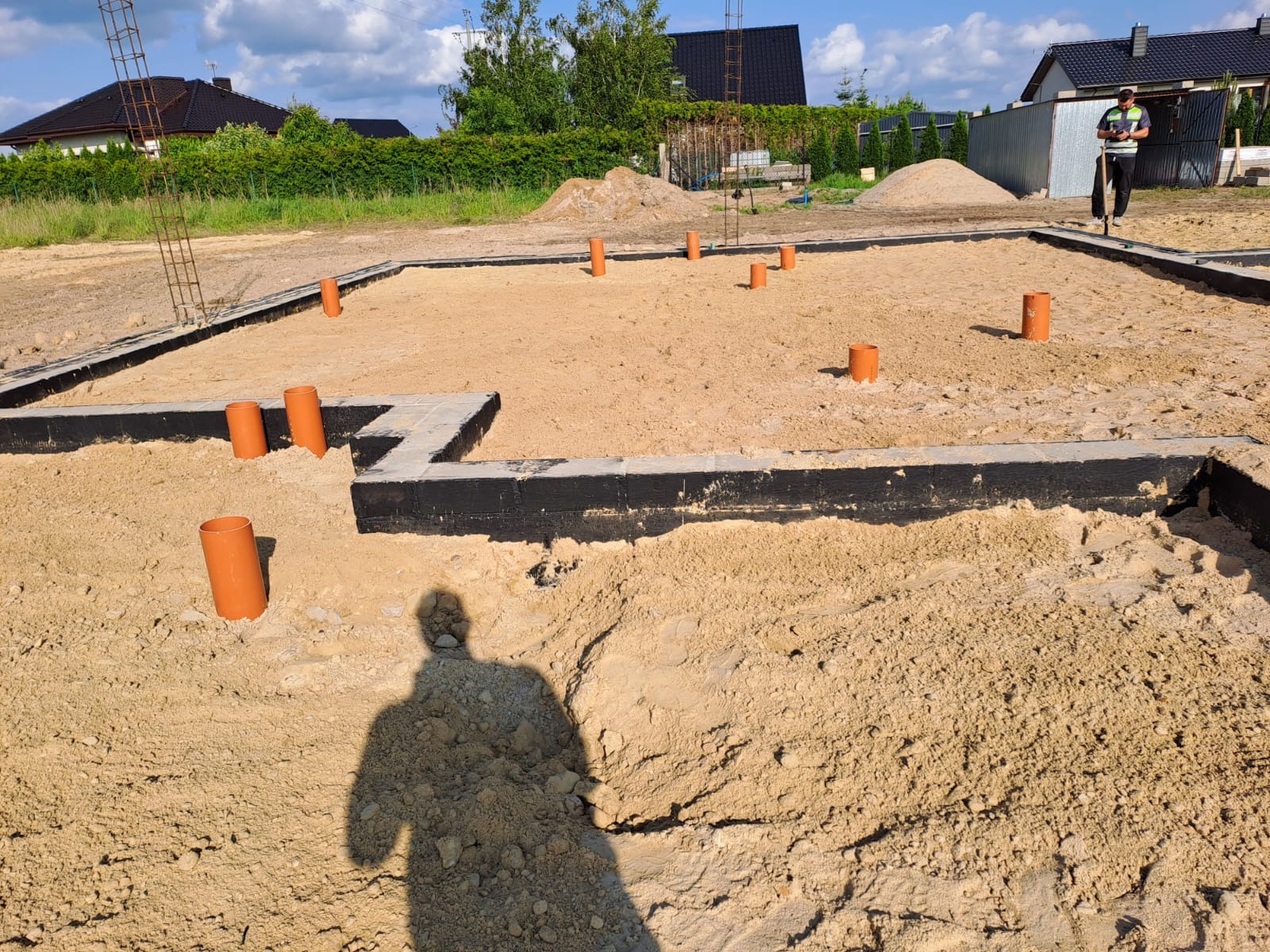 Przygotowanie fundamentów pod budynek z wystającymi rurami kanalizacyjnymi, zabezpieczone hydroizolacją, teren wyrównany piaskiem, widoczny cień osoby.