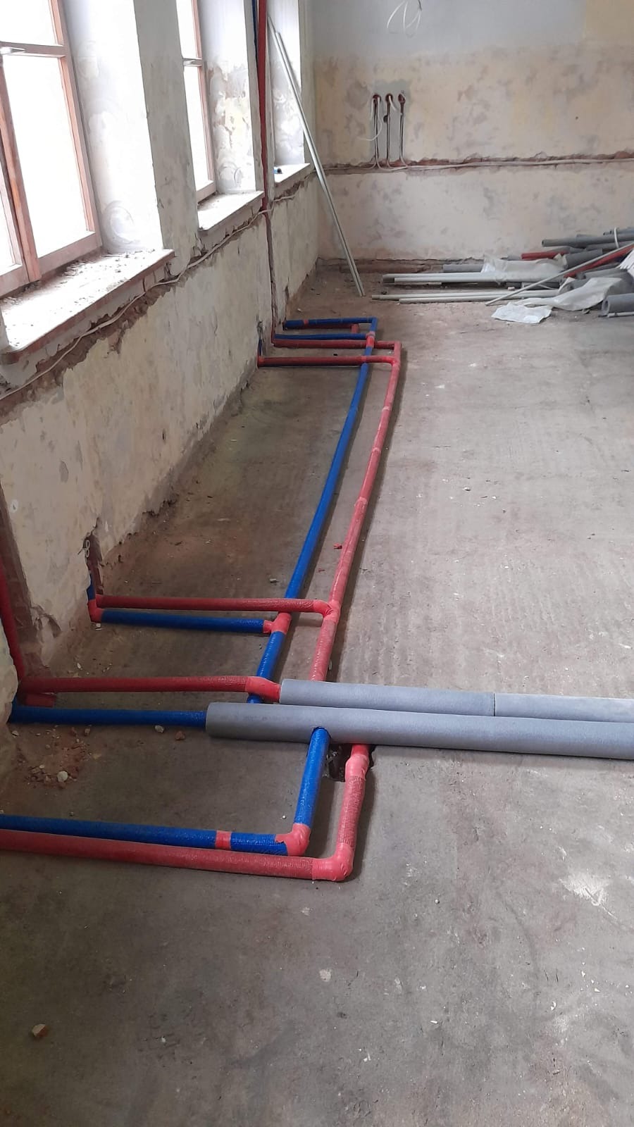 Instalacja wodna w trakcie montażu: czerwone i niebieskie rury prowadzone wzdłuż ściany, szare rury izolowane leżące na podłodze. Surowy stan wykończenia.