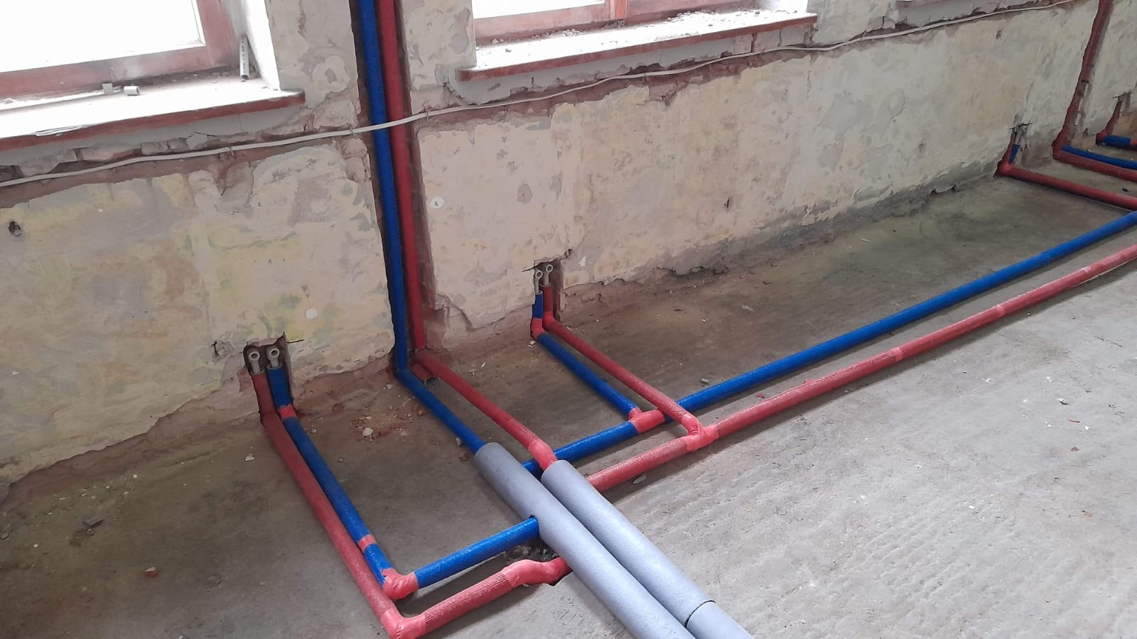 Instalacja ogrzewania podłogowego w trakcie montażu: niebieskie i czerwone rury ułożone na surowym betonie, wychodzące ze ściany, widoczne okno w tle.