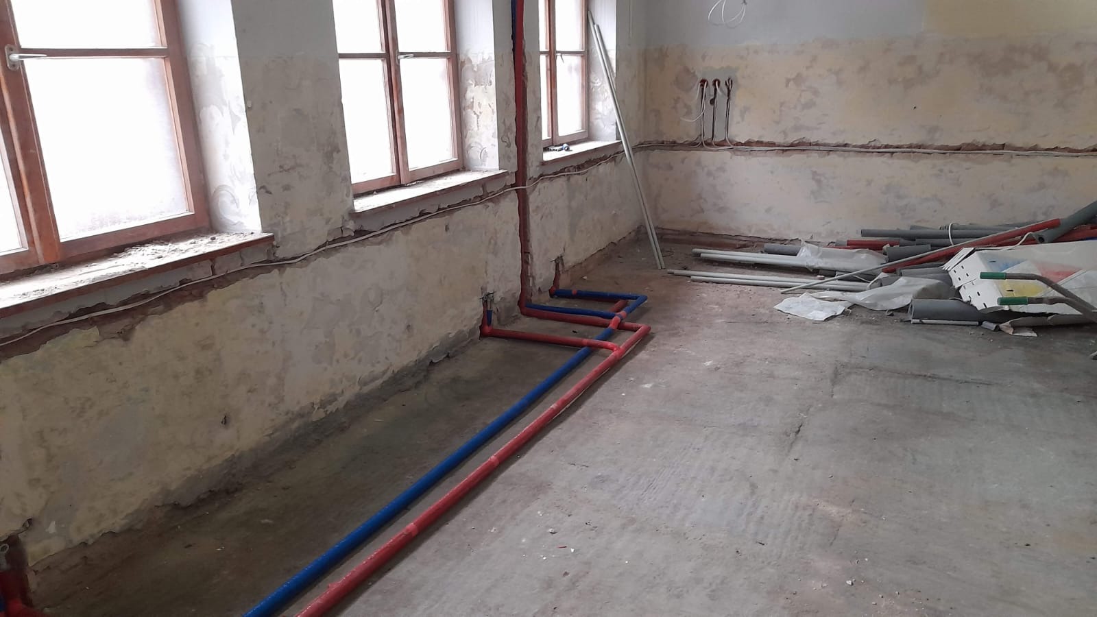 Instalacja wodna z rur czerwonych i niebieskich w surowym wnętrzu z oknami i fragmentami instalacji elektrycznej. Widoczne materiały budowlane.