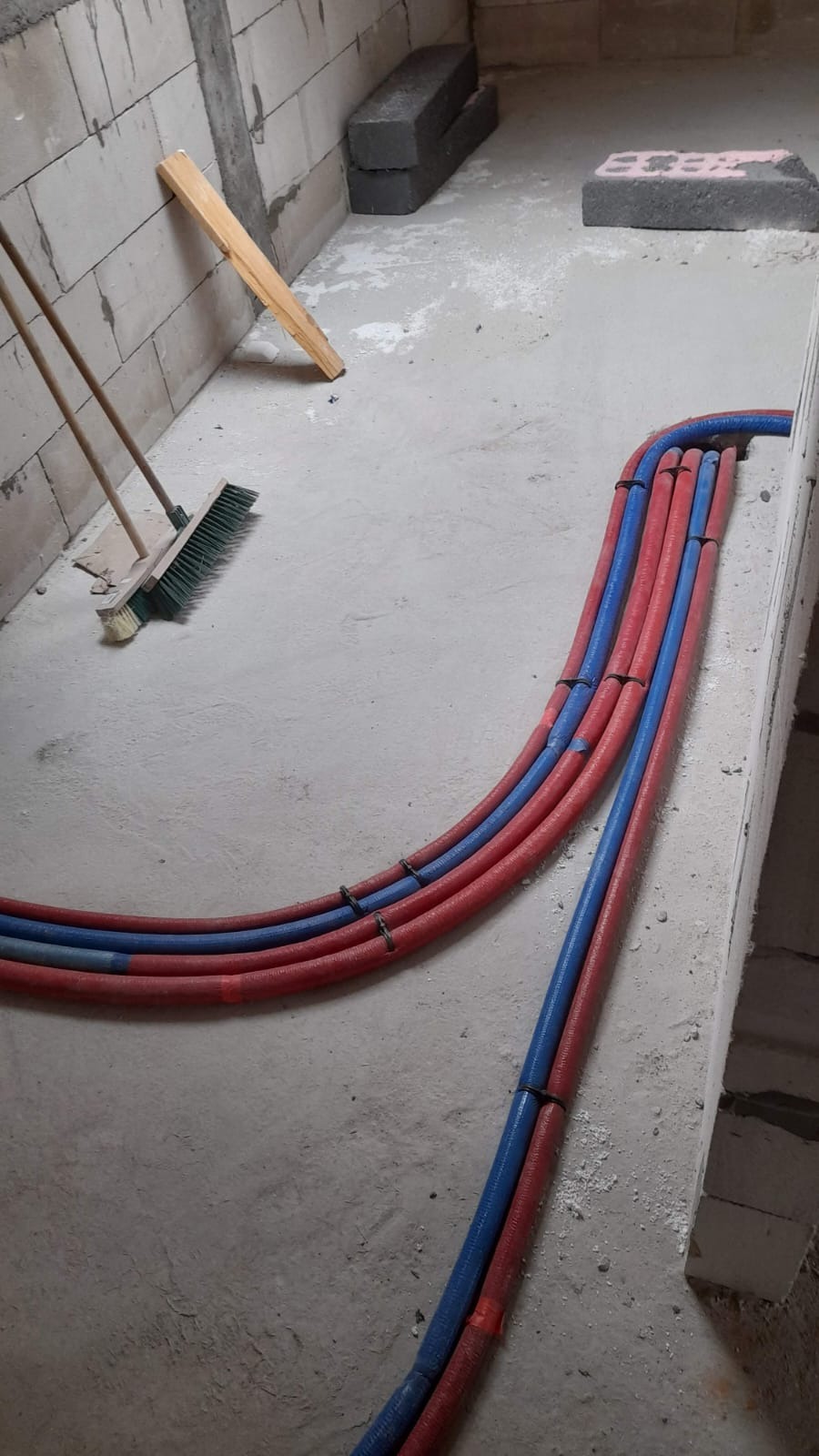 Ułożone, czerwono-niebieskie rury ogrzewania podłogowego w surowym pomieszczeniu, przymocowane czarnymi opaskami. W tle cegły i narzędzia.