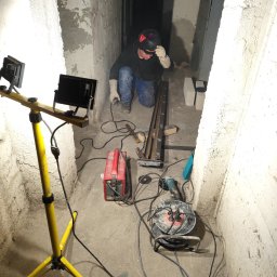 AEM SYSTEM Ewa Seńków-Dziedzic - Pracownik w masce spawalniczej klęczy w wąskim korytarzu z surowymi ścianami, przygotowując metalowe elementy pod oświetleniem halogenowym, obok czerwonej spawarki i szlifierki kątowej.