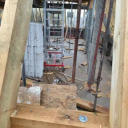 Wnętrze budynku w trakcie budowy, widoczne drewniane elementy konstrukcyjne, podpory stalowe, rusztowanie na kółkach, oraz stos pustaków na podmokłej podłodze