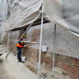 AEM SYSTEM Ewa Seńków-Dziedzic - Pracownik w kasku i kamizelce na rusztowaniu, wyrównuje zaprawą fragment ceglanej ściany budynku, zabezpieczonego siatką ochronną.