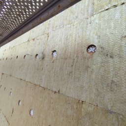 AEM SYSTEM Ewa Seńków-Dziedzic - Ściana budynku w trakcie docieplania, widoczne płyty izolacyjne z otworami wypełnionymi białymi kołkami mocującymi, ujęcie z rusztowania.