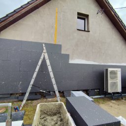 AEM SYSTEM Ewa Seńków-Dziedzic - Dom w trakcie nakładania ciemnoszarego styropianu na elewację, widoczna drabina, zaprawa w pojemniku i klimatyzator na zewnątrz budynku.
