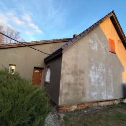 AEM SYSTEM Ewa Seńków-Dziedzic - Dom w trakcie prac elewacyjnych: część ściany pokryta szarym tynkiem, druga część z widocznymi śladami po wcześniejszych pracach, okno zabezpieczone folią, widoczny fragment dachu z ciemną dachówką...