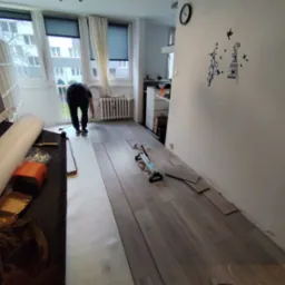 Układanie paneli podłogowych w pokoju, widoczne narzędzia, częściowo ułożona podłoga i osoba pracująca przy grzejniku.