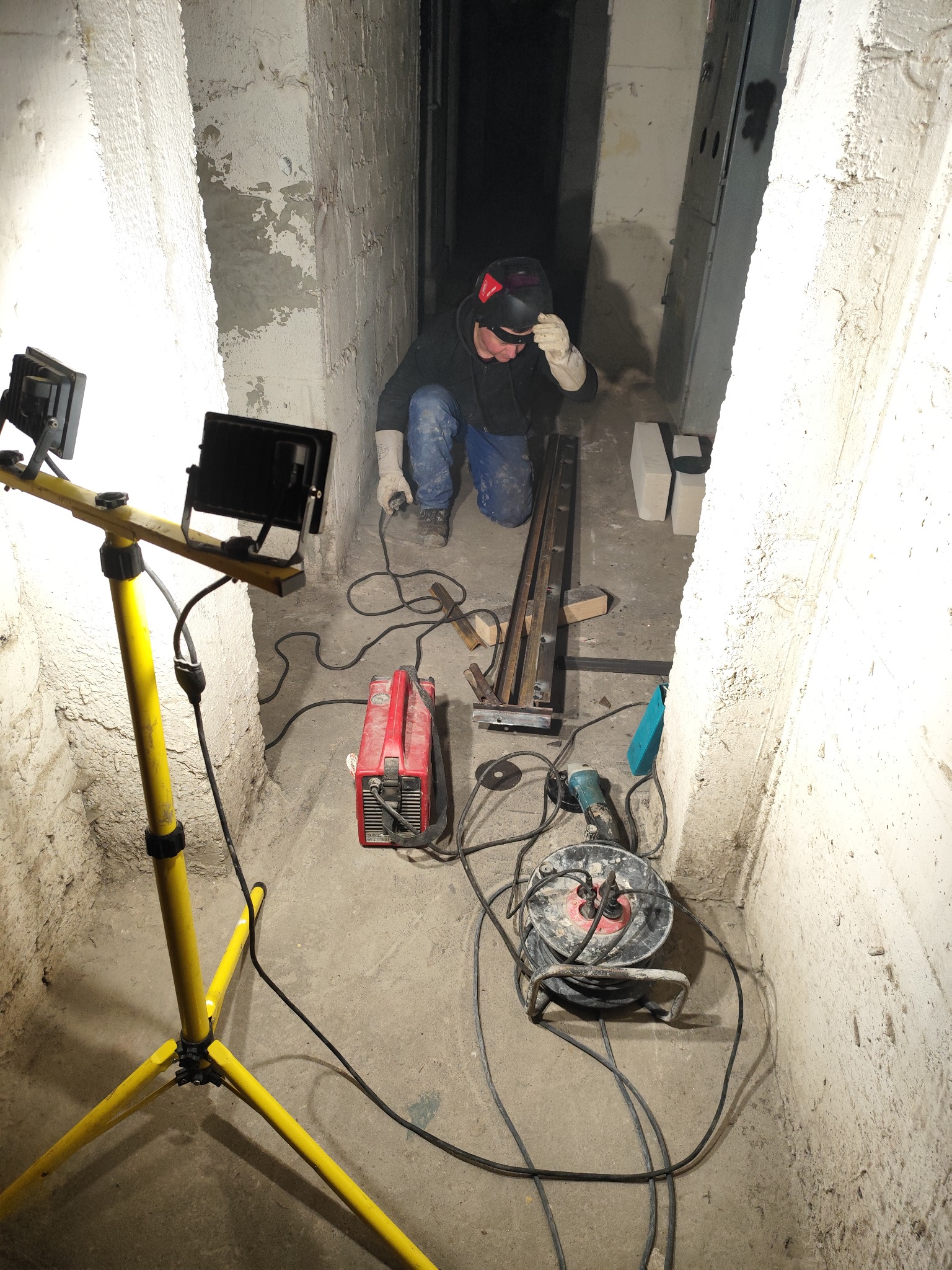 Pracownik w masce spawalniczej klęczy w wąskim korytarzu z surowymi ścianami, przygotowując metalowe elementy pod oświetleniem halogenowym, obok czerwonej spawarki i szlifierki kątowej.