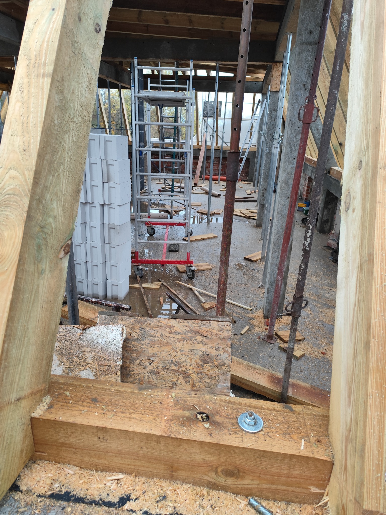 Wnętrze budynku w trakcie budowy, widoczne drewniane elementy konstrukcyjne, podpory stalowe, rusztowanie na kółkach, oraz stos pustaków na podmokłej podłodze