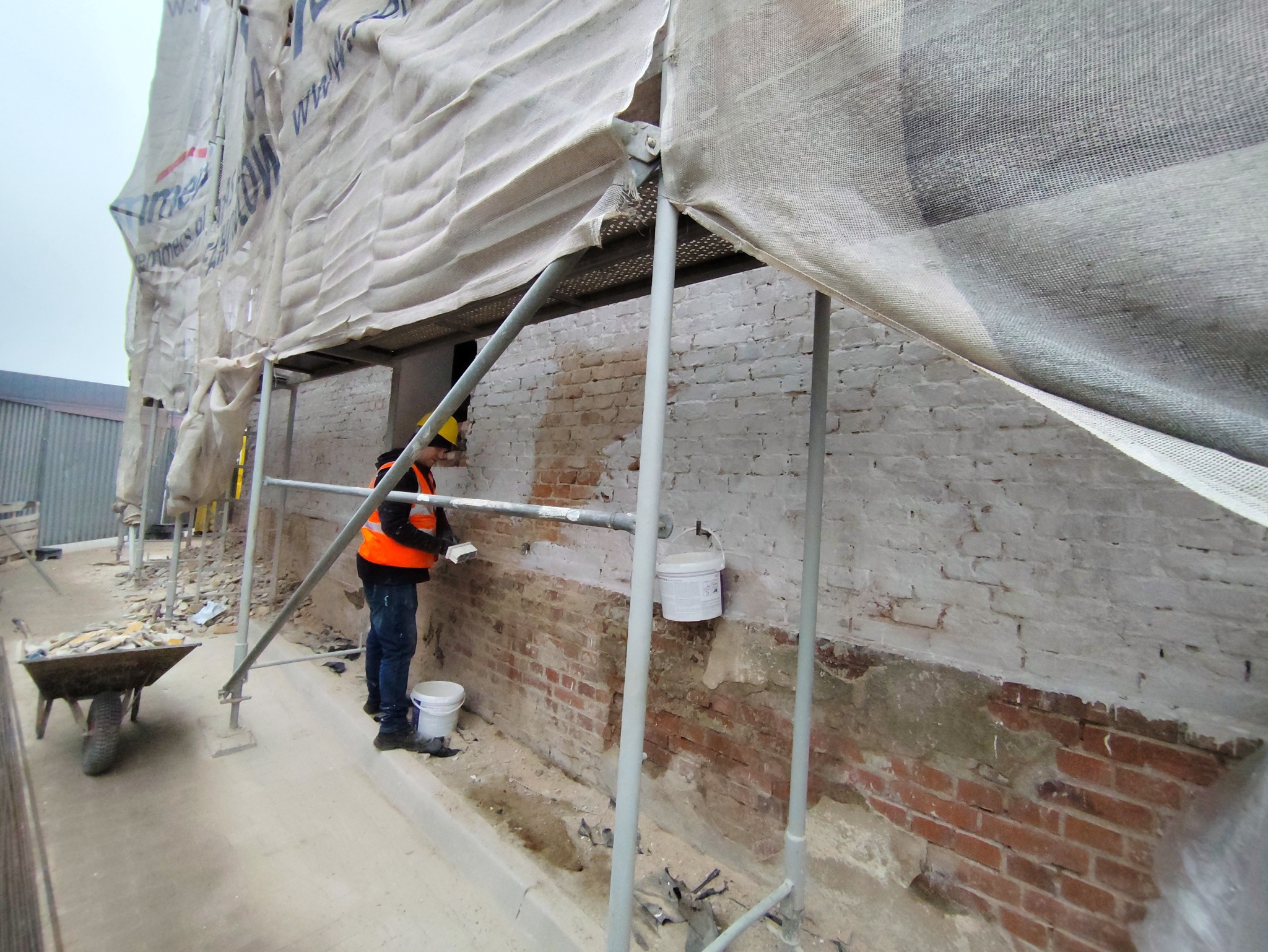Pracownik w kasku i kamizelce na rusztowaniu, wyrównuje zaprawą fragment ceglanej ściany budynku, zabezpieczonego siatką ochronną.