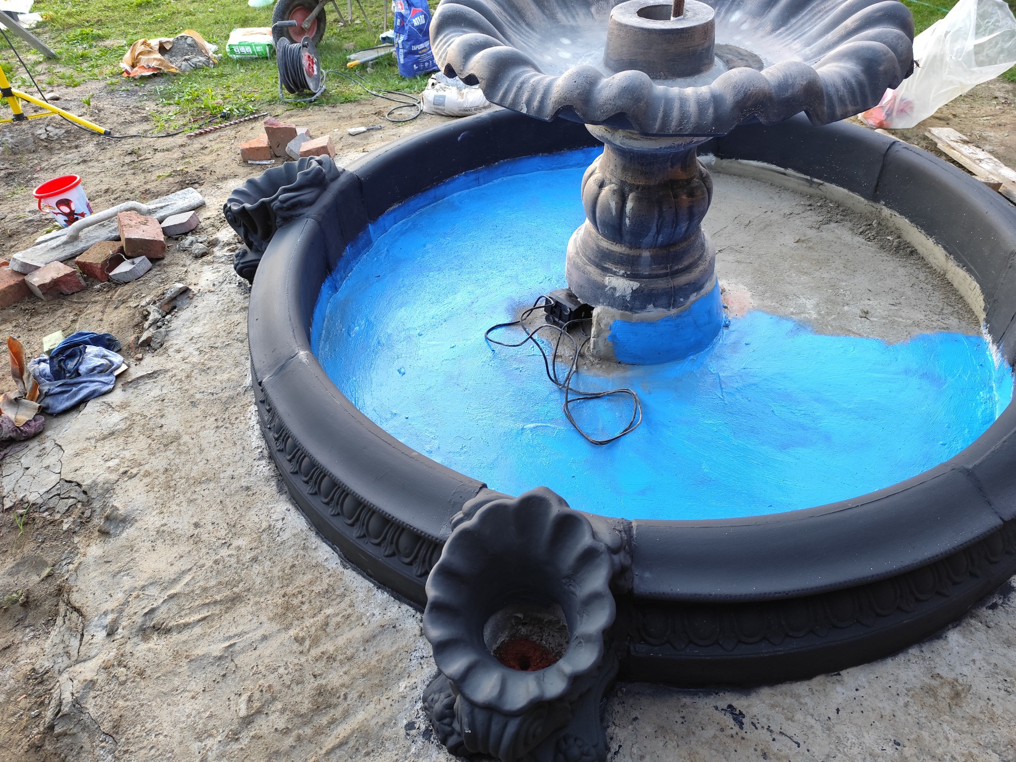 Okrągła fontanna ogrodowa z czarną obudową i świeżo pomalowanym na niebiesko wnętrzem zbiornika na wodę, widoczne narzędzia i materiały budowlane w tle.
