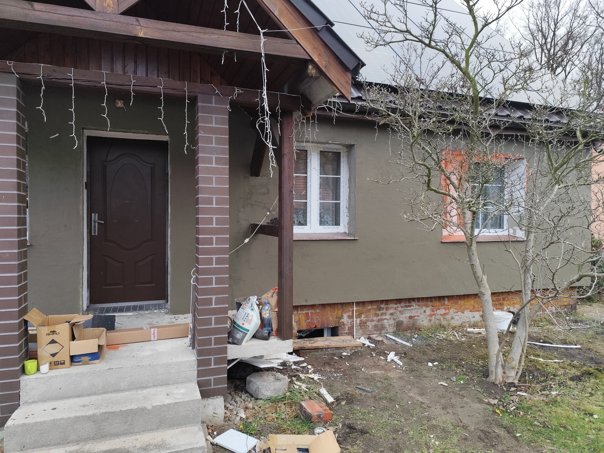 Dom jednorodzinny w trakcie prac elewacyjnych: świeżo otynkowana ściana w odcieniach szarości, widoczne worki z zaprawą, resztki materiałów budowlanych na ziemi i dekoracyjne lampki zwisające...