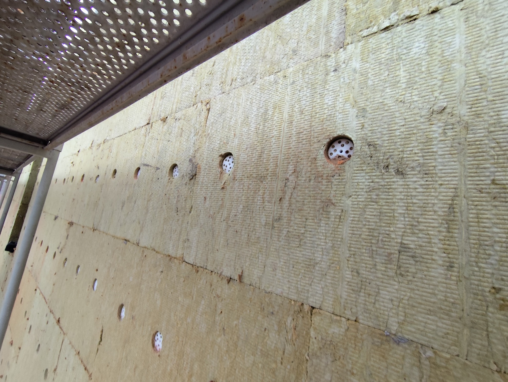 Ściana budynku w trakcie docieplania, widoczne płyty izolacyjne z otworami wypełnionymi białymi kołkami mocującymi, ujęcie z rusztowania.