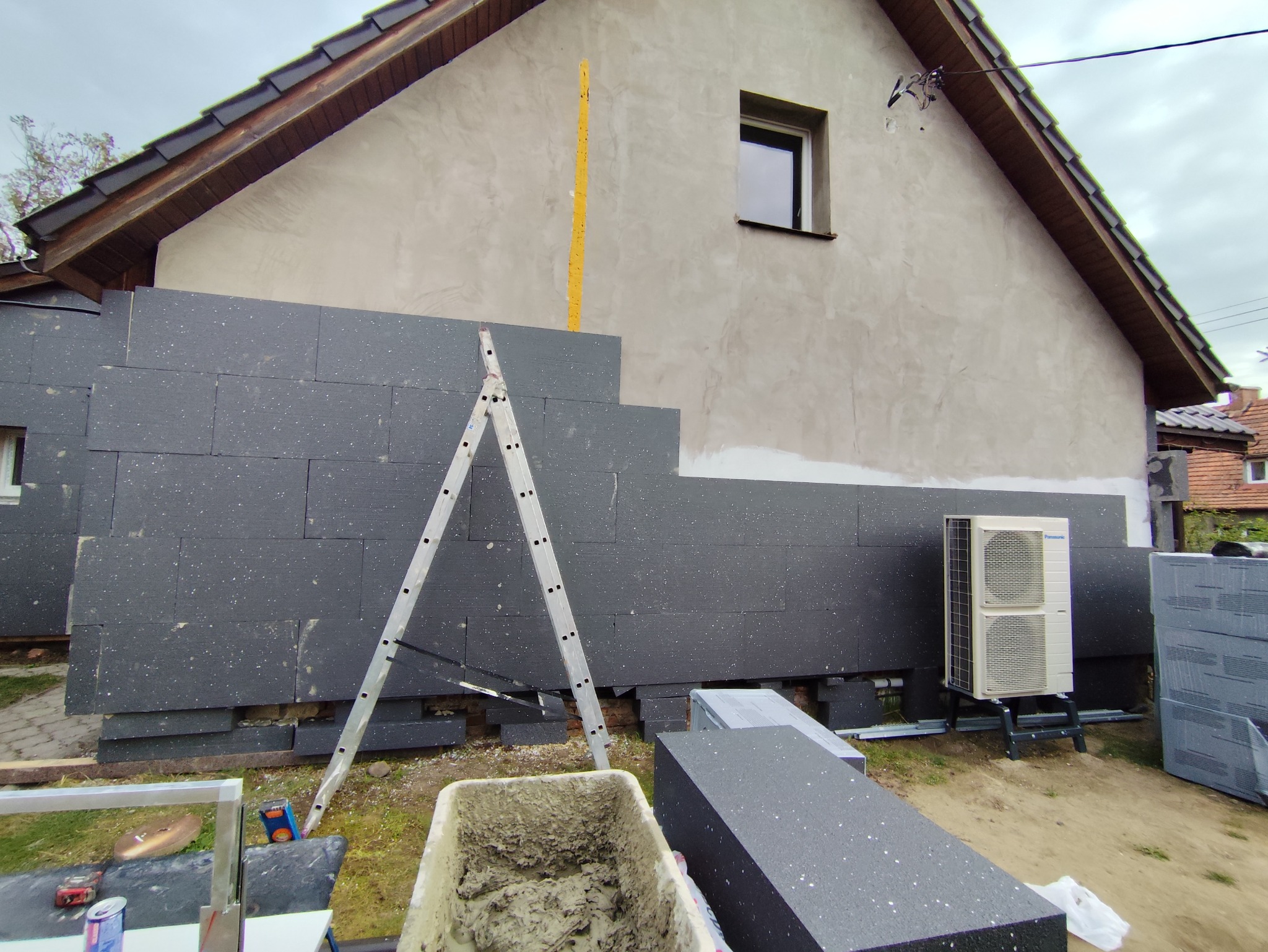Dom w trakcie nakładania ciemnoszarego styropianu na elewację, widoczna drabina, zaprawa w pojemniku i klimatyzator na zewnątrz budynku.