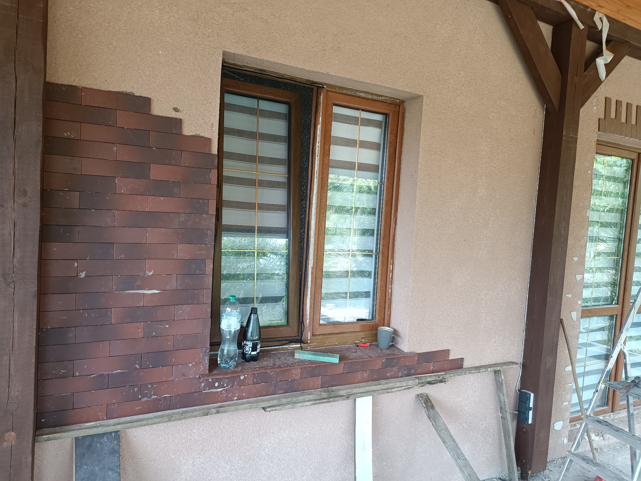 Fragment elewacji budynku z częściowo ułożoną okładziną z cegieł klinkierowych w odcieniach czerwieni i brązu, widoczne okno z drewnianą ramą, butelki z napojami na parapecie i elementy rusztowania.