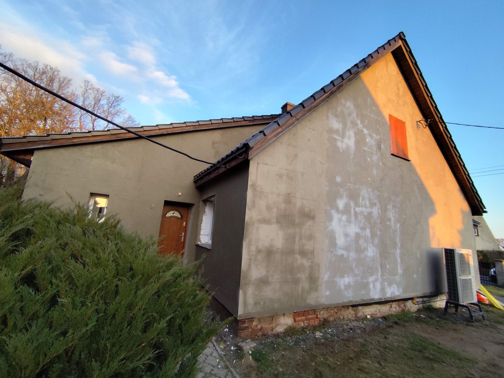 Dom w trakcie prac elewacyjnych: część ściany pokryta szarym tynkiem, druga część z widocznymi śladami po wcześniejszych pracach, okno zabezpieczone folią, widoczny fragment dachu z ciemną dachówką...