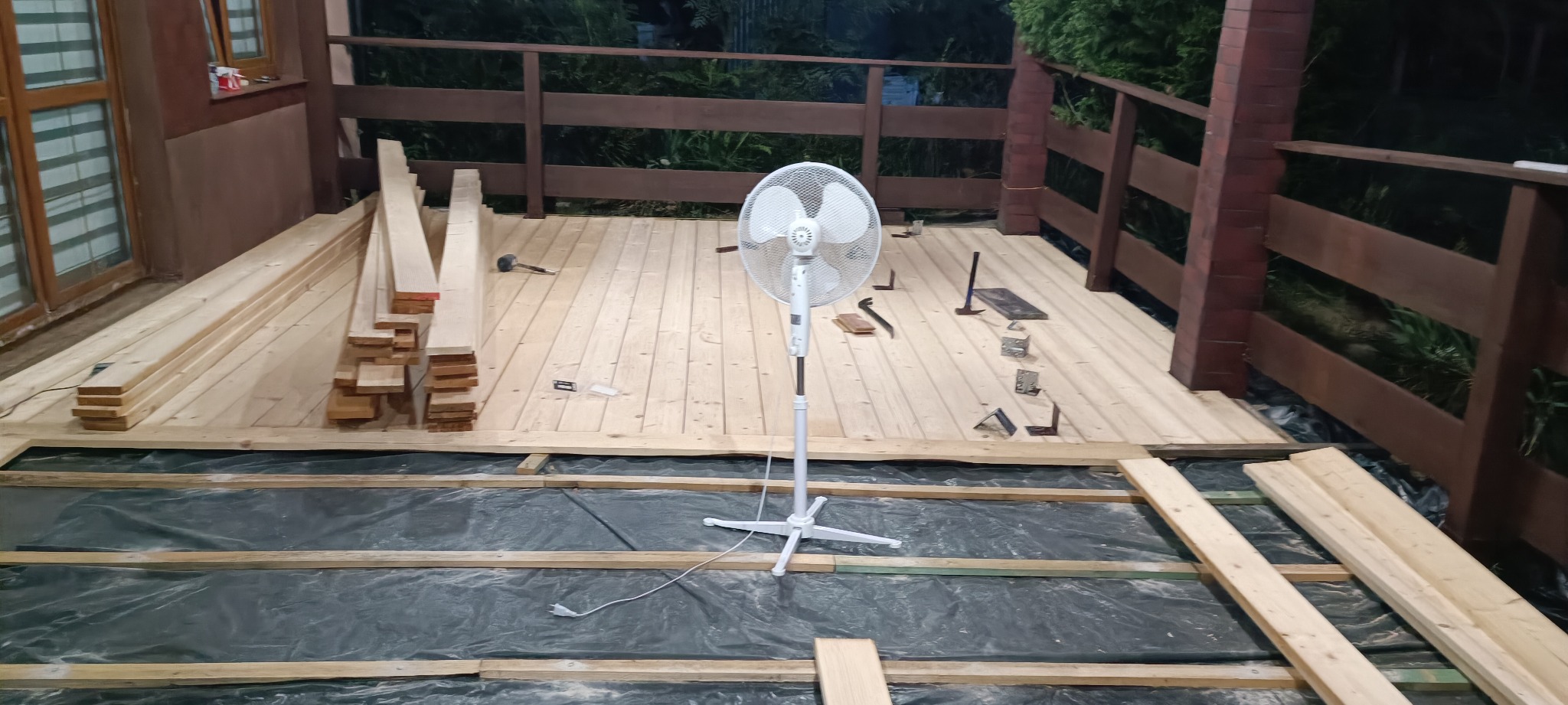 Budowa drewnianego tarasu: widok na częściowo ułożone deski, wentylator stojący, narzędzia i materiały budowlane na podłożu z czarnej folii.