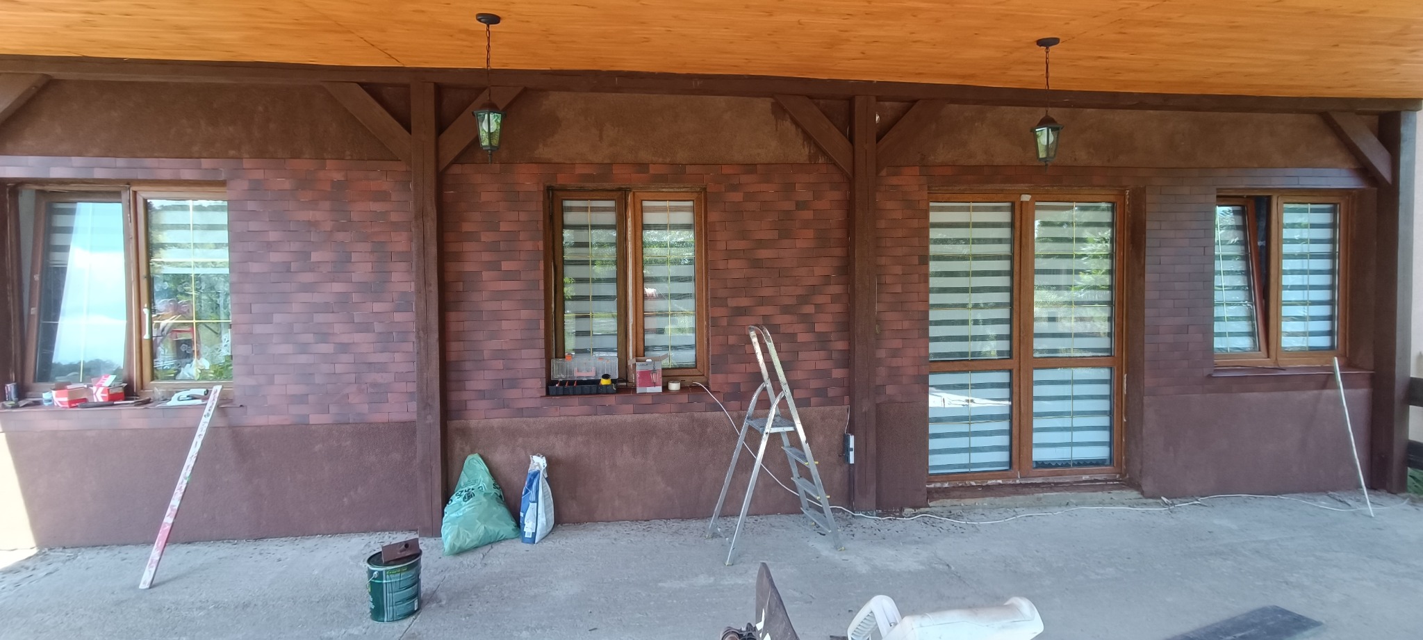 Elewacja domu wykończona cegłą klinkierową, widoczne okna z roletami, drewniana konstrukcja podtrzymująca dach, drabina, worki z materiałami budowlanymi i puszka po farbie.