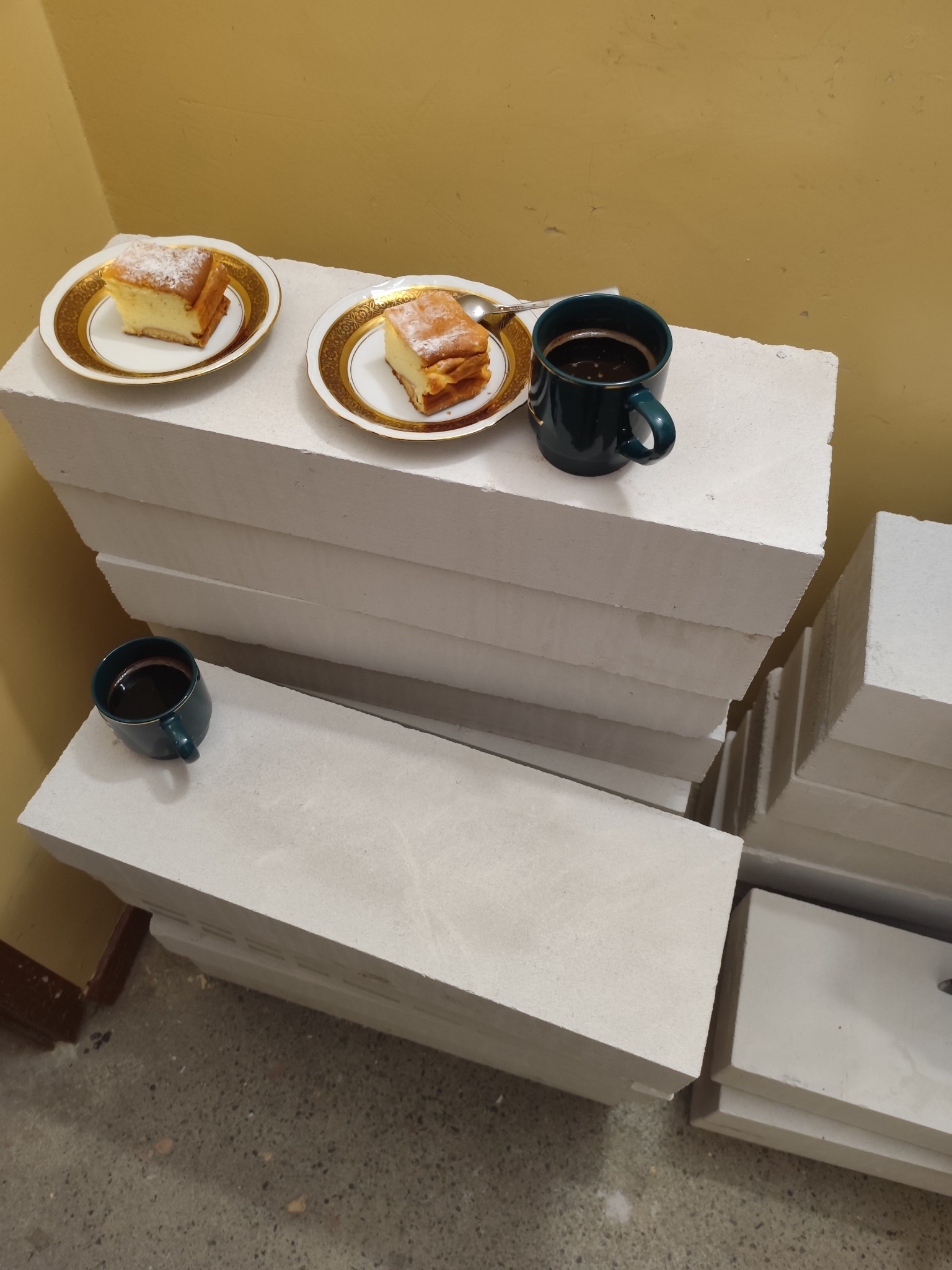 Dwa kawałki sernika na talerzykach z pozłacanym brzegiem i dwie filiżanki czarnej kawy na stosie białych bloczków z betonu komórkowego.