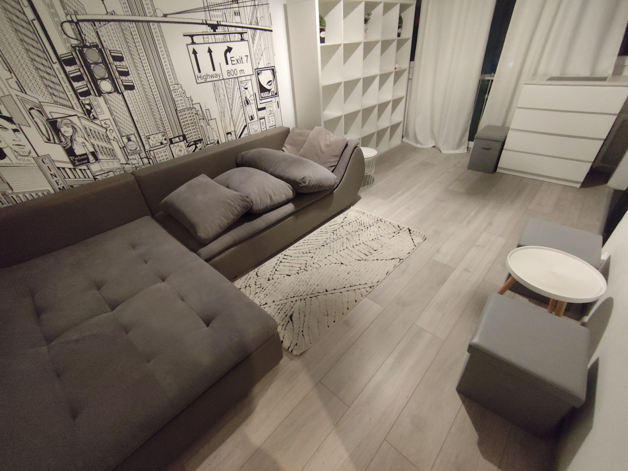 Szary narożnik z poduszkami na tle ściany z rysunkiem miasta, dywan z geometrycznym wzorem i białe meble w nowoczesnym salonie.