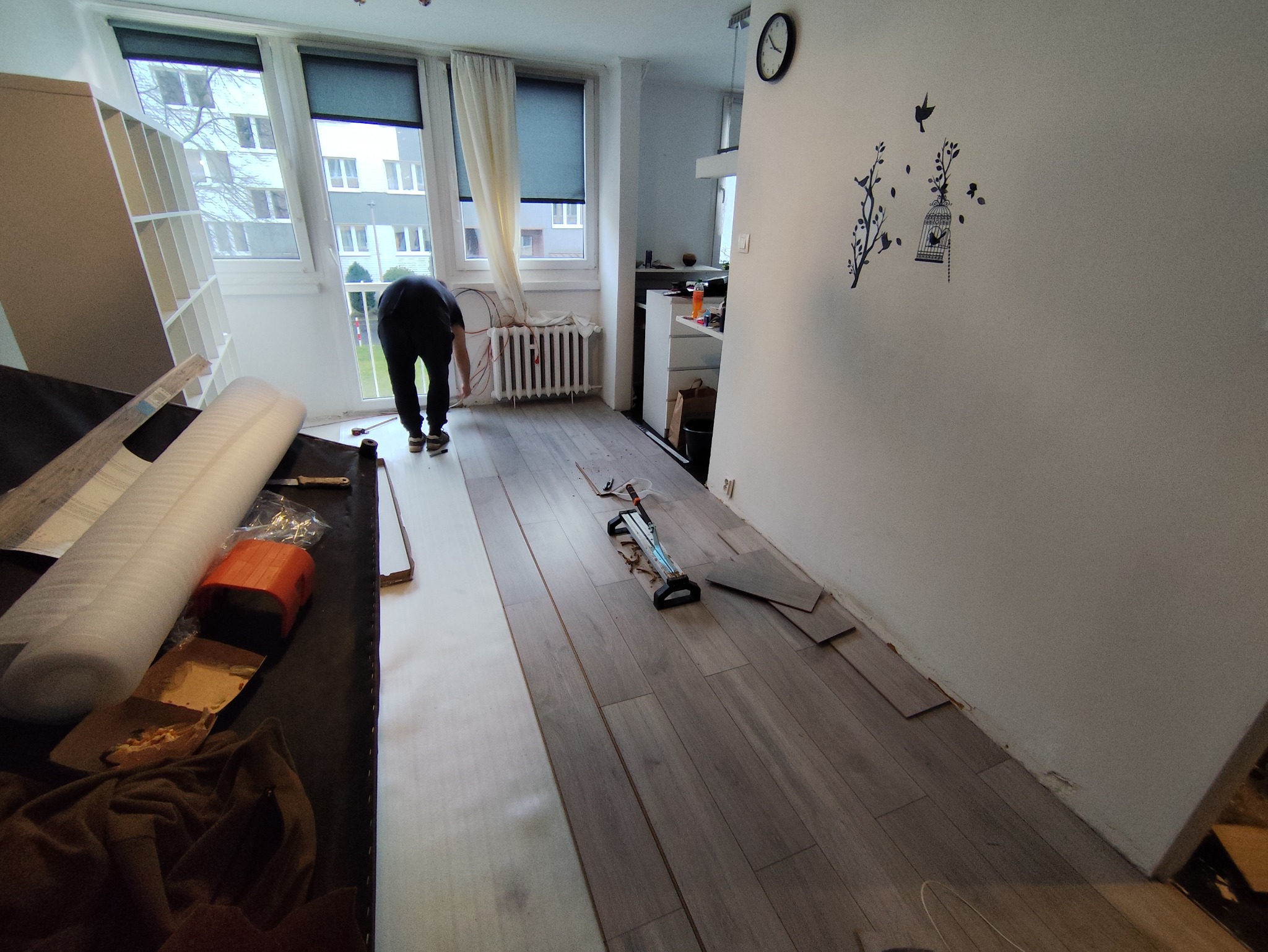 Układanie paneli podłogowych w pokoju, widoczne narzędzia, częściowo ułożona podłoga i osoba pracująca przy grzejniku.