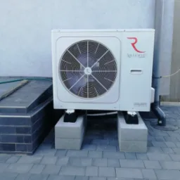 Biała jednostka zewnętrzna urządzenia grzewczego marki Rotenso Aquami Series, umieszczona na betonowych bloczkach na szarej kostce brukowej, z widoczną rurą spustową i fragmentem schodów.