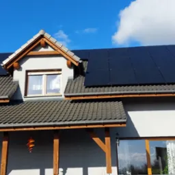 Dom jednorodzinny z panelami fotowoltaicznymi na dachu, widok z dołu na elewację i dach, błękitne niebo z chmurami w tle.