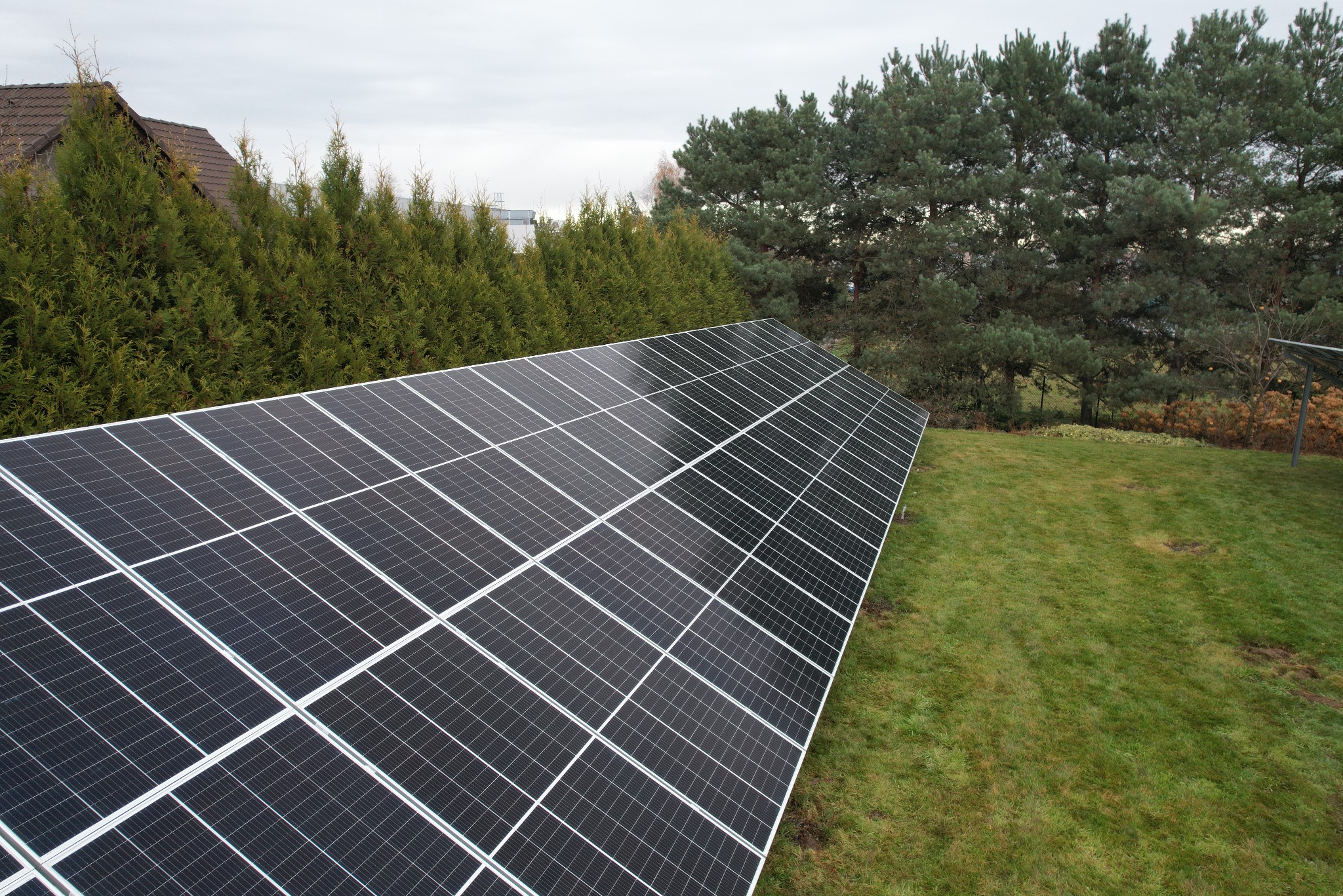 Instalacja paneli słonecznych na trawniku, widok z góry, w tle iglaki i dach domu.