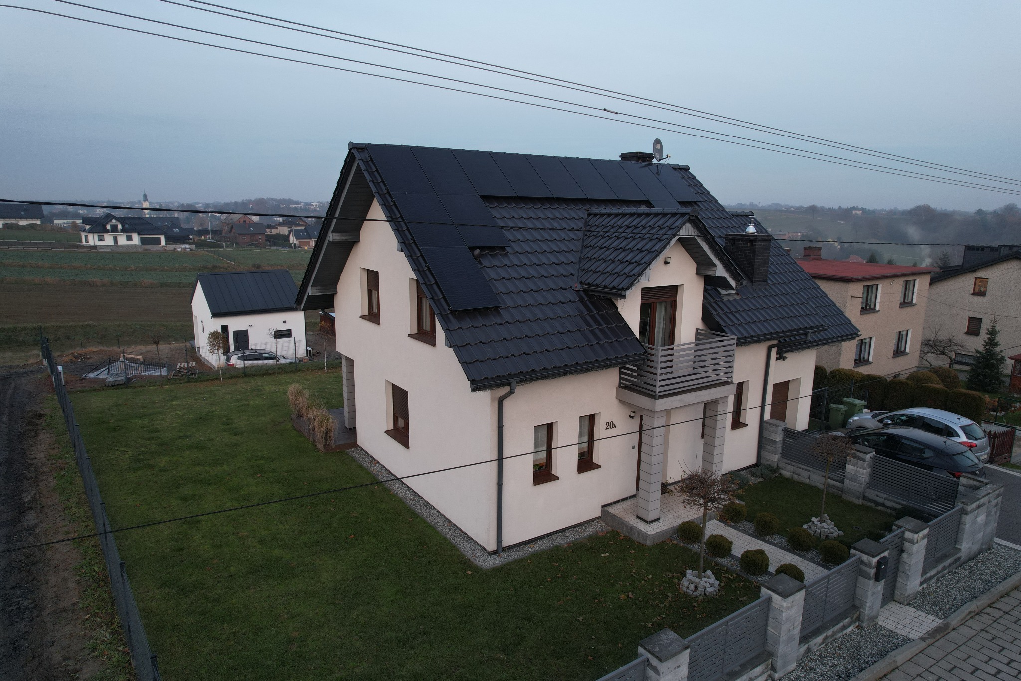 Nowoczesny dom jednorodzinny z ciemną dachówką i panelami słonecznymi na dachu, widok z góry na ogród i okoliczną zabudowę w pochmurny dzień.