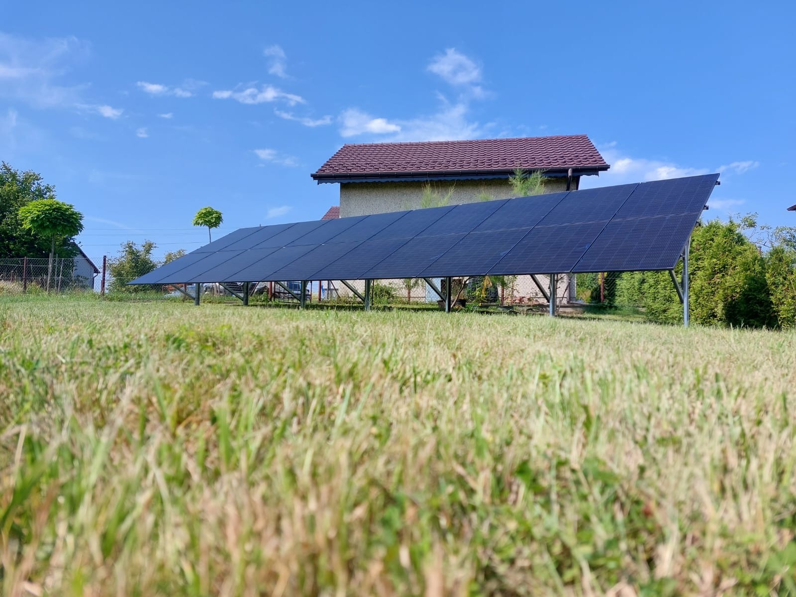 Rząd paneli słonecznych na metalowej konstrukcji, ustawiony na trawiastym wzgórzu przed domem z czerwoną dachówką, widok z poziomu trawy.