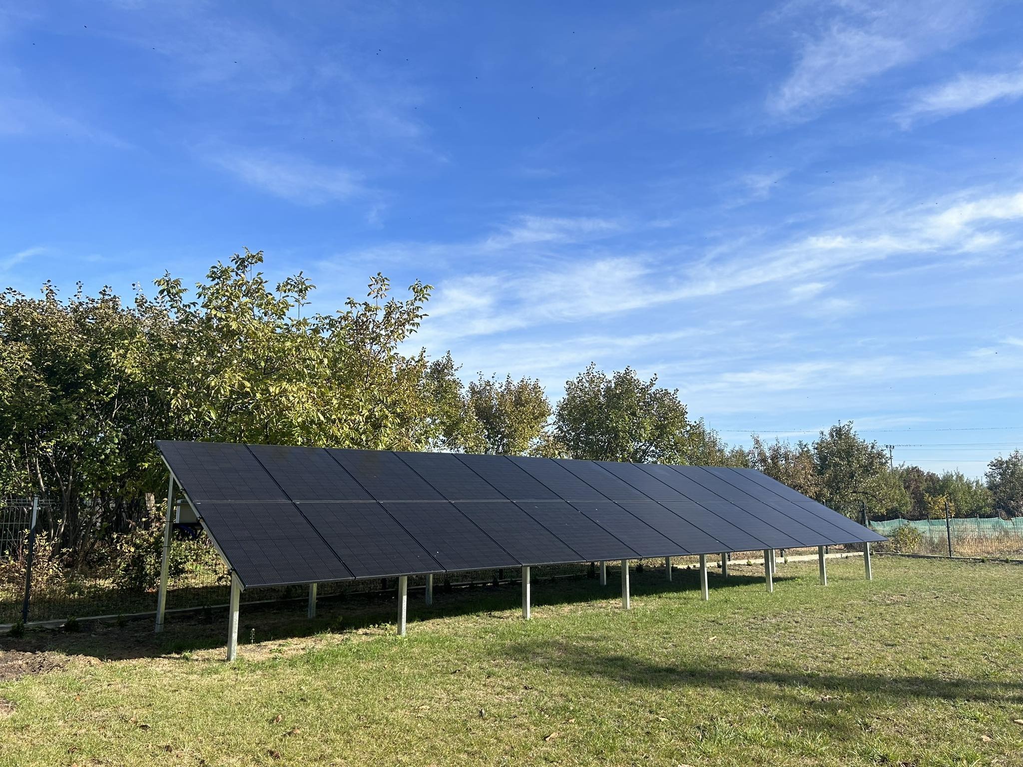 Rząd czarnych paneli słonecznych na metalowych podporach ustawiony na trawiastym terenie w słoneczny dzień z błękitnym niebem i drzewami w tle.