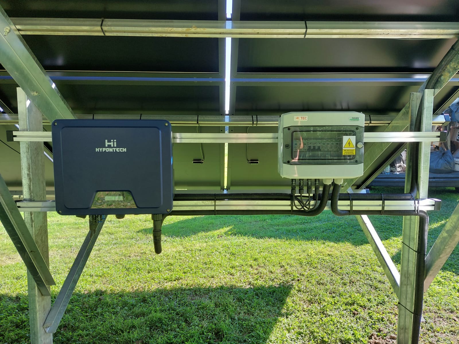 Instalacja fotowoltaiczna: widok od spodu na konstrukcję wsporczą z zamontowanym inwerterem Hyponttech oraz skrzynką przyłączeniową na tle zielonego trawnika.
