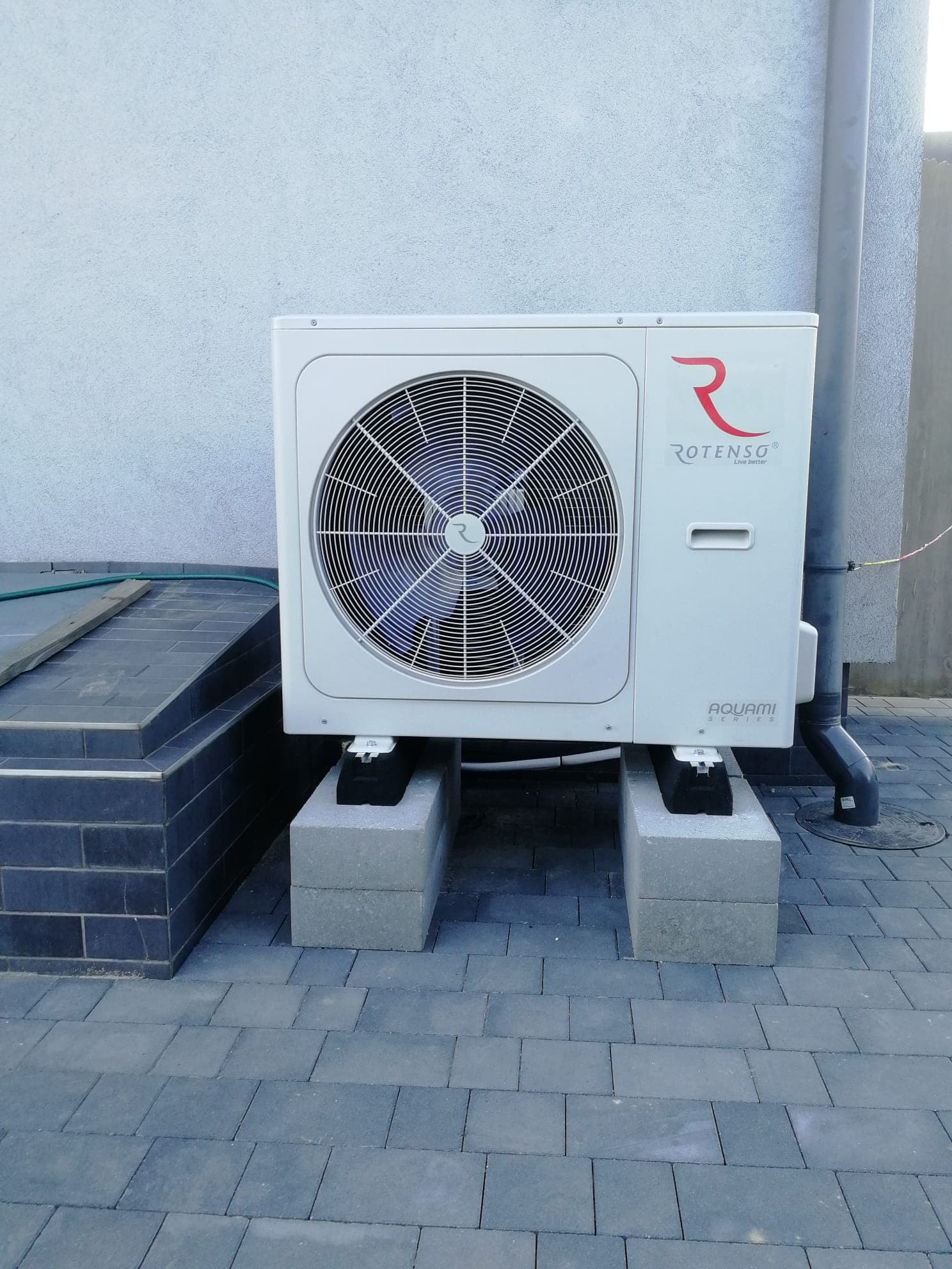 Biała jednostka zewnętrzna urządzenia grzewczego marki Rotenso Aquami Series, umieszczona na betonowych bloczkach na szarej kostce brukowej, z widoczną rurą spustową i fragmentem schodów.