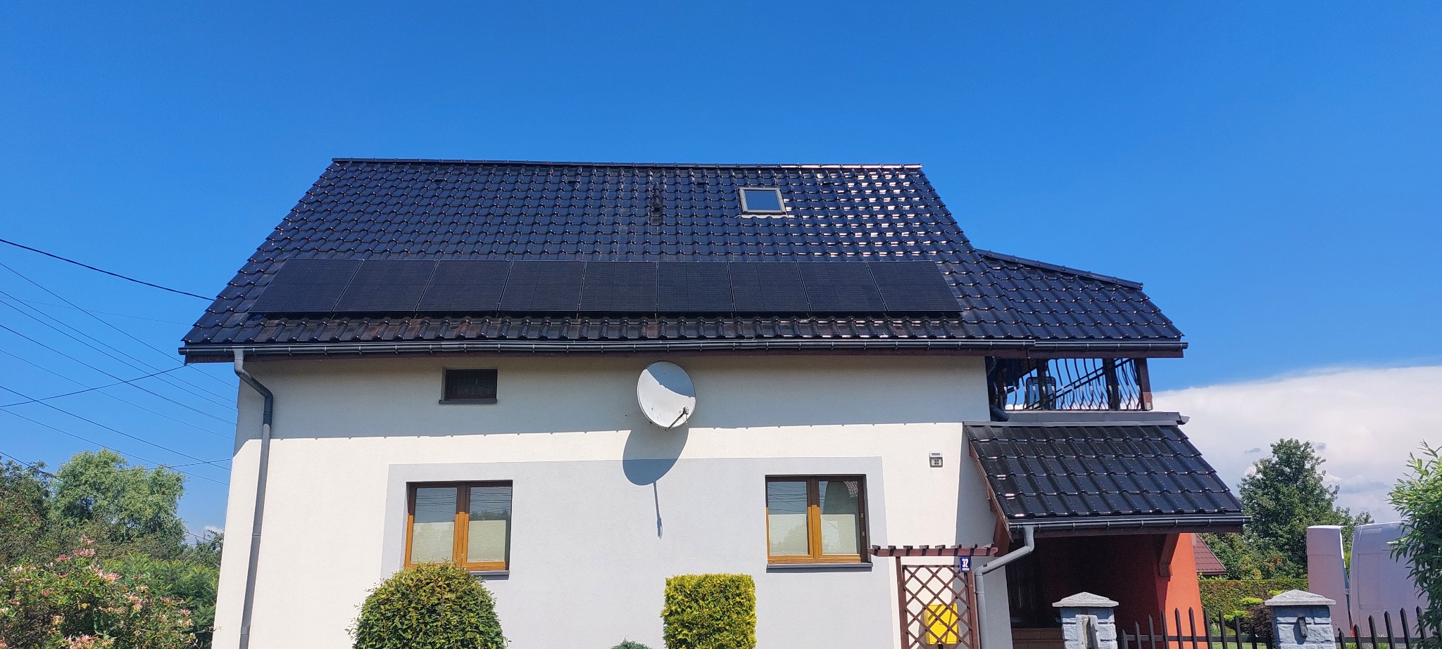 Dwukondygnacyjny dom z ciemną dachówką i panelami słonecznymi na dachu, antena satelitarna na elewacji, błękitne niebo w tle.