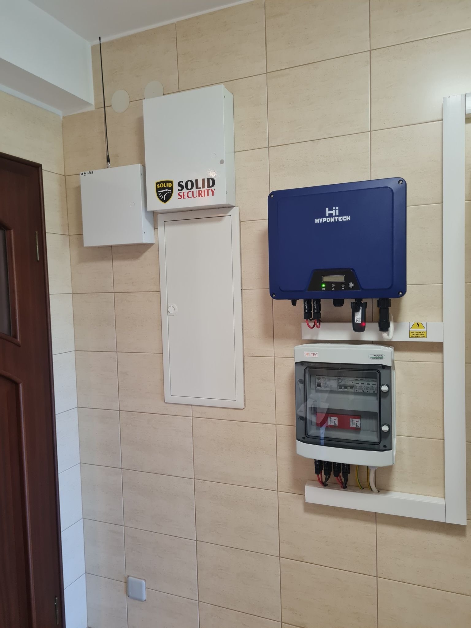 Wnętrze pomieszczenia z zainstalowanym niebieskim inwerterem fotowoltaicznym marki Hi HYPONTECH, podłączonym do skrzynki z bezpiecznikami, zamontowanymi na kafelkowanej ścianie obok brązowych drzwi.